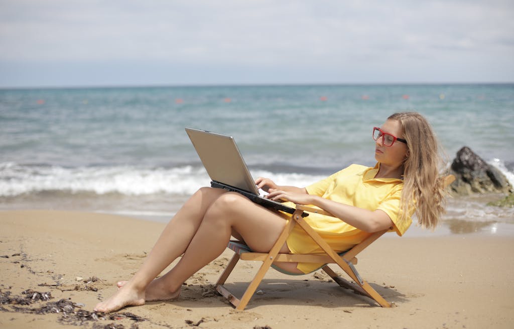 A woman with blond hair wearing a yellow top relaxes on a beach chair while using a laptop by the seashore.